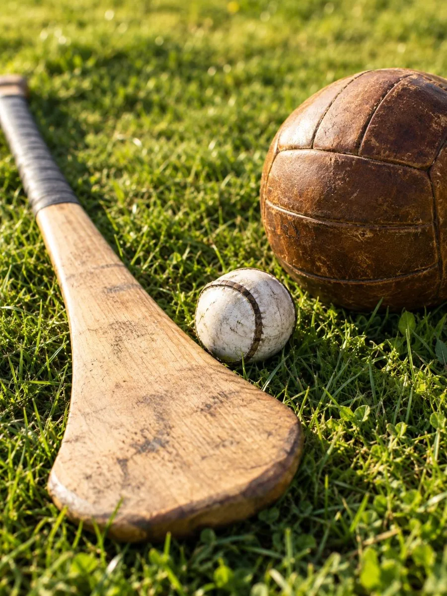 Ein hölzerner Hurleystock, ein kleiner weißer Sliotar und ein Leder-Gaelic-Football auf sonnenbeschienener Wiese